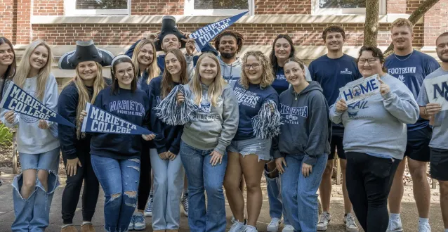 16 Marietta College students stand together, all smiling, wearing navy blue, gray, and white Marietta College attire, holding Marietta College flags and signs